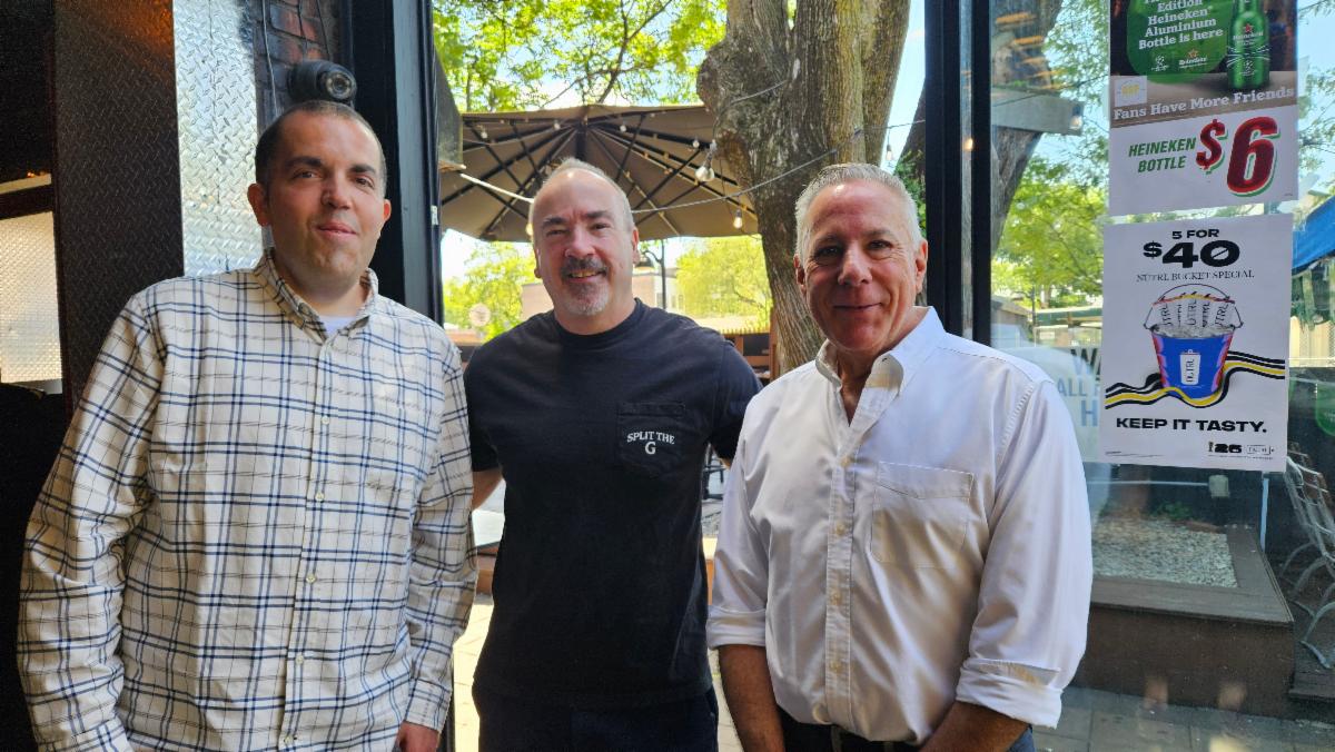 Three men standing together inside a cafe near a window with outdoor seating and promotional signs visible