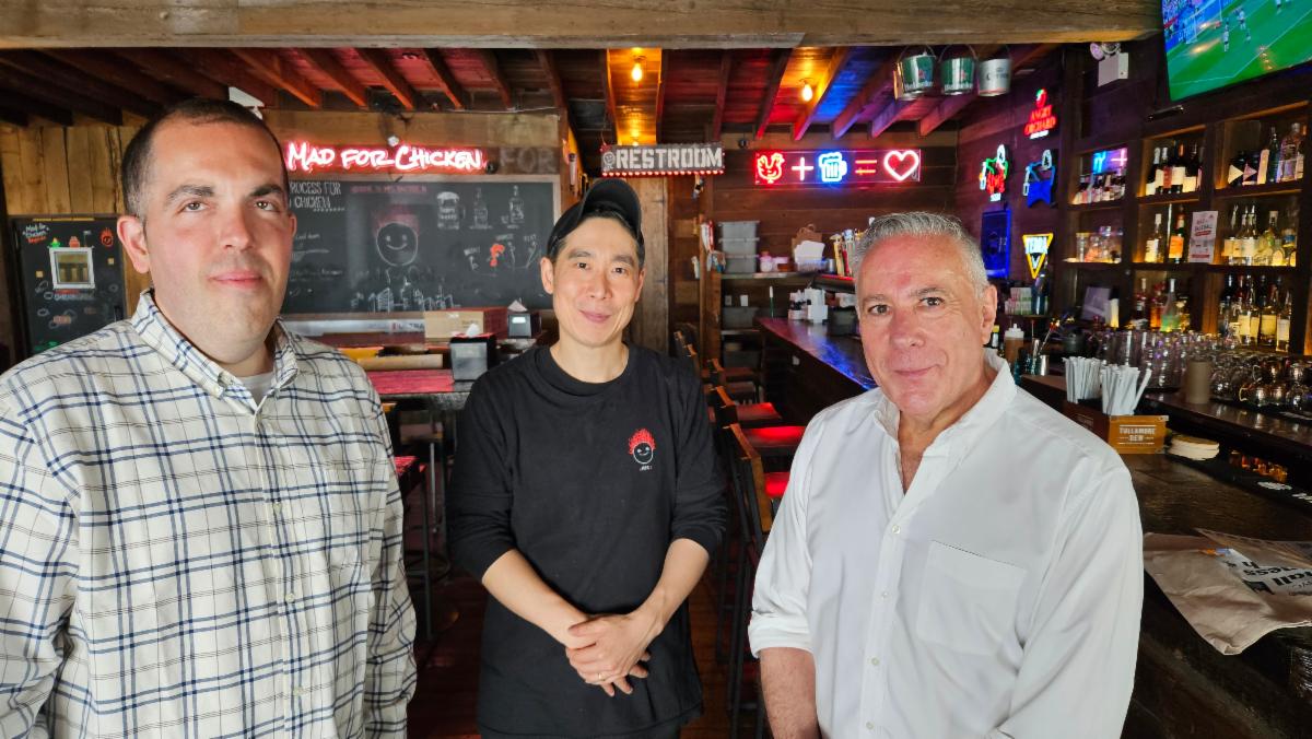 Three men standing inside a cozy bar with wooden beams, neon signs, and a well-stocked liquor shelf in the background