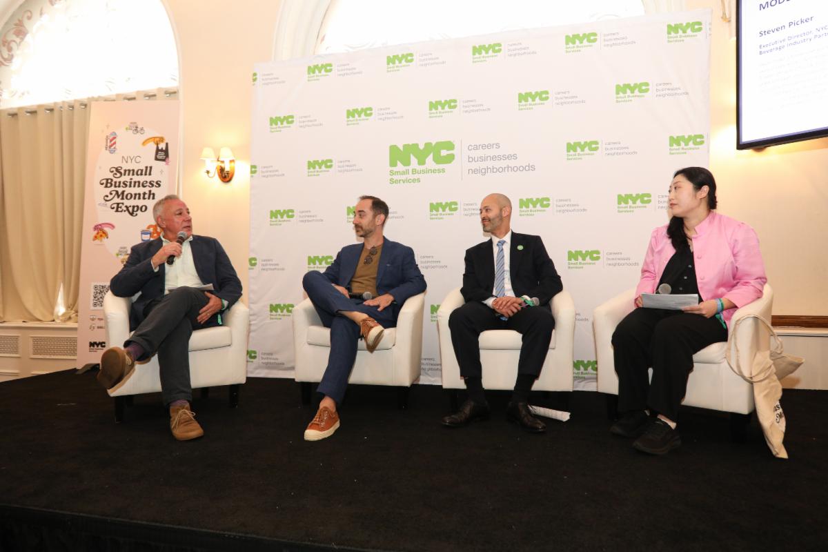 Four panelists seated on stage at NYC Small Business Month Expo discussing business topics with branded backdrop behind them.