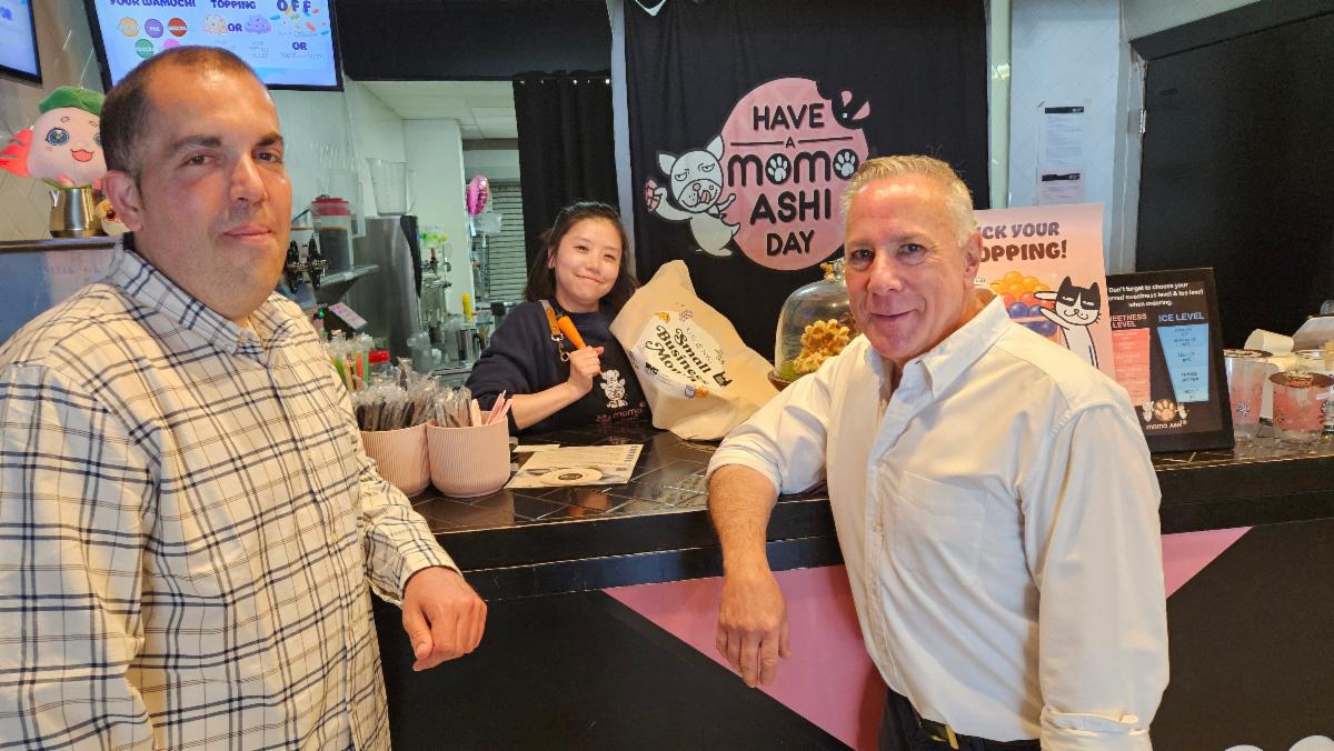 Two customers standing at a counter in a brightly lit food shop with a sign reading 'Have a Mommi Ashi Day' in the background