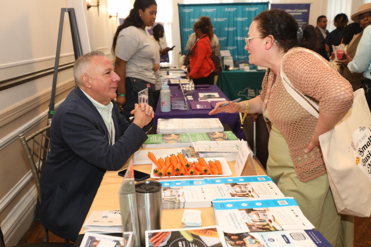 Man seated at a booth handing out brochures and promotional items to a woman leaning forward to ask questions at a busy indoor event.