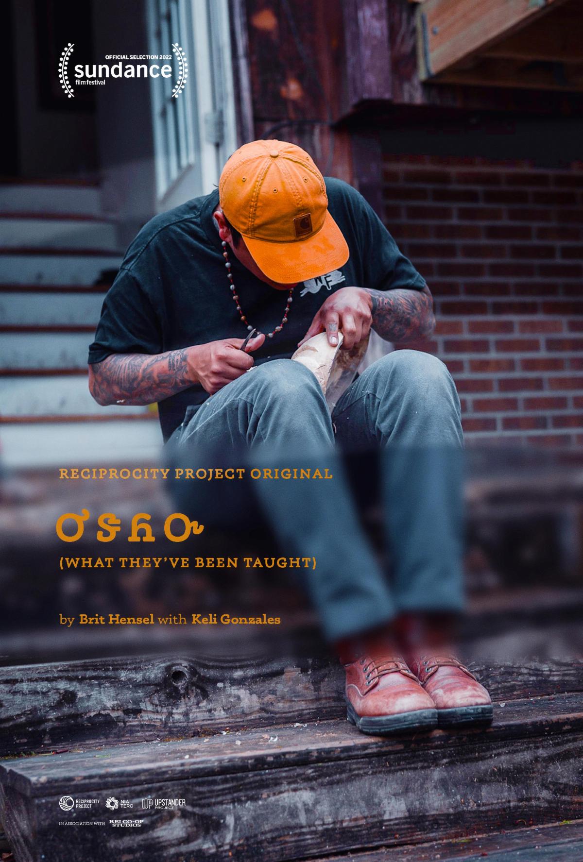 A person in jeans and an orange hat with tattooed arms sits on a stoop outside of a brick building and is hunched over carving something out of stone. 
