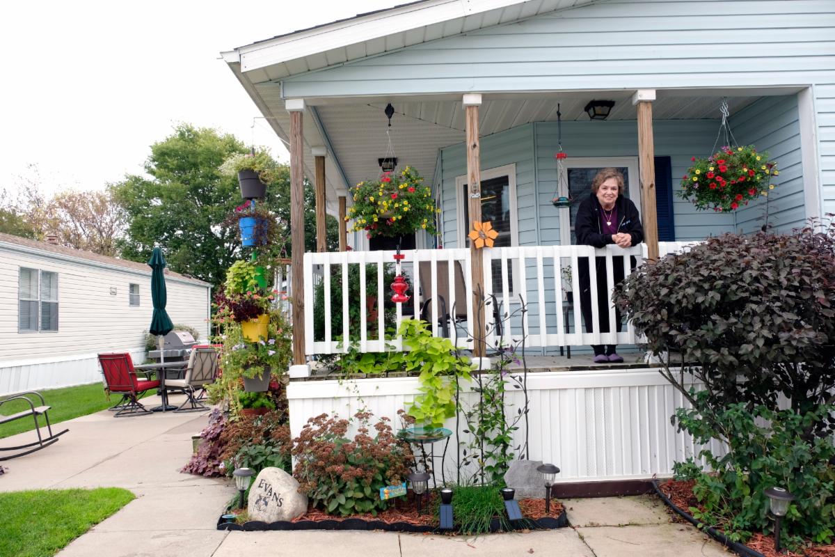 An image of a woman who appears to be in her 70s standing on her front porch and smiling. It is a small cozy looking home with pretty little gardens around it. She leans over the railing of the porch and smiles.