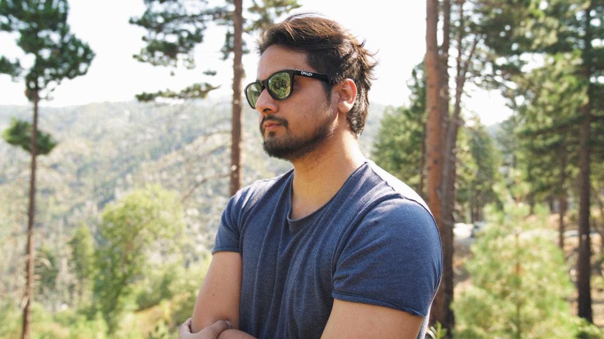 Rajee is shown in profile with his arms crossed in front of a forest. He has sunglasses, short dark hair and a dark beard. He wears a blue t-shirt.