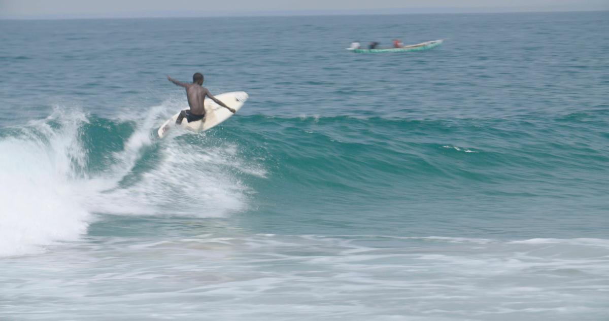 A black man is surfing a small wave on a cream colored surfboard. A small boat is motoring past in the background.