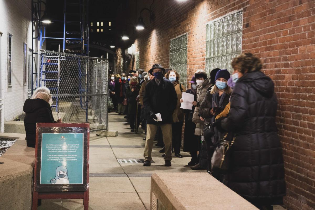 A long line of people in winter coats and face masks stretches alongside a brick building at nighttime, outside a theater waiting to see a film. 