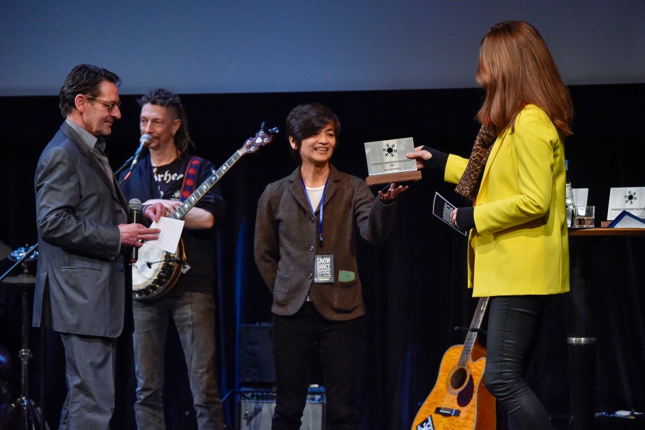 The filmmaker, Des, onstage receiving an award. She is wearing a dark suit and the woman handing her the award is wearing a bright yellow jacket. To the filmmaker's left is a man holding a microphone and directly behind them is a man holding a guitar.