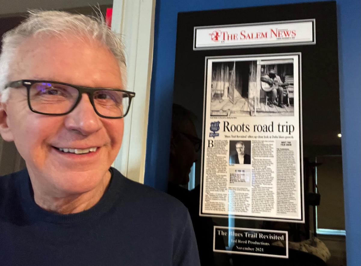 A white man with short gray hair and glasses smiles in front of a framed newspaper article from The Salem News. The headline reads: Roots Road Trip.