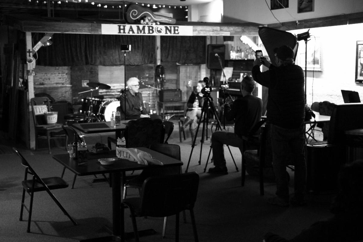 A black and white photograph of an interview being held in an old blues club. A woman interviews man on a stage while in the forefront cameramen are recording it. At the top of the rickety stage is a sign that reads "Hambone".