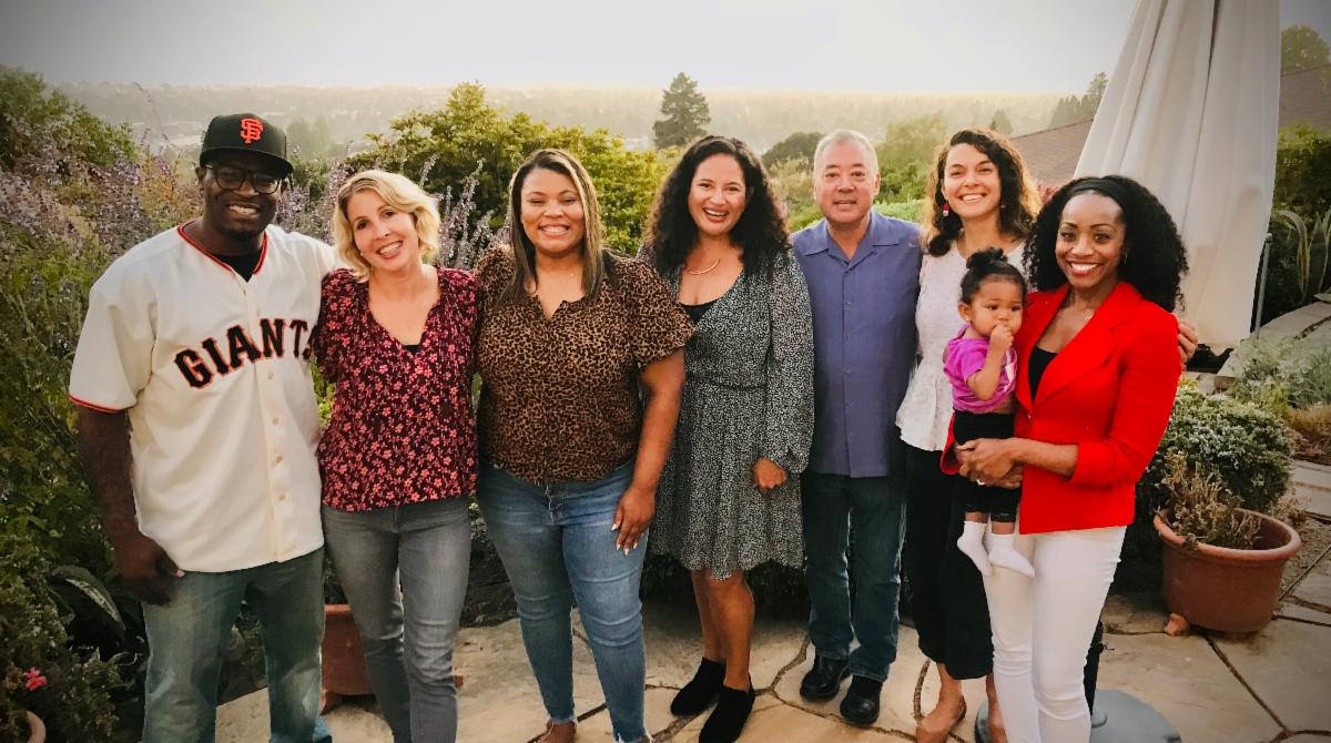 A group of five women and two men are gathered standing together smiling and posing for a picture outside on a patio on a sunny day. They are smiling, and the woman on the far right is holding a small child. 
