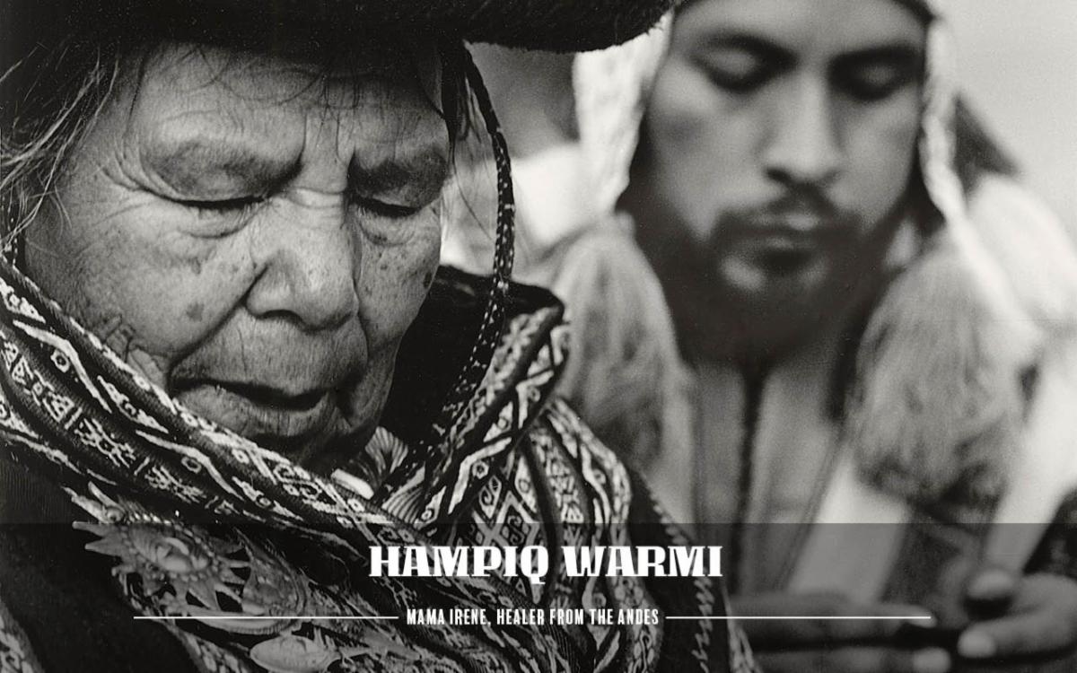 A black and white photo. An old woman of indigenous descent stands in forefront with her eyes closed and appears to be speaking in mid-prayer. She wears a hat and a patterned scarf. In the backdrop is a younger man also with eyes closed.