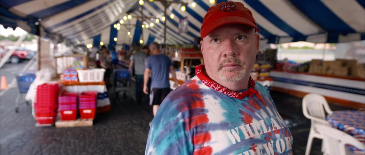 A heavyset middle-aged man standing in under a large tent where fireworks are sold. He wears a red baseball cap, a red bandana around his neck and a blue tie-dye t-shirt.