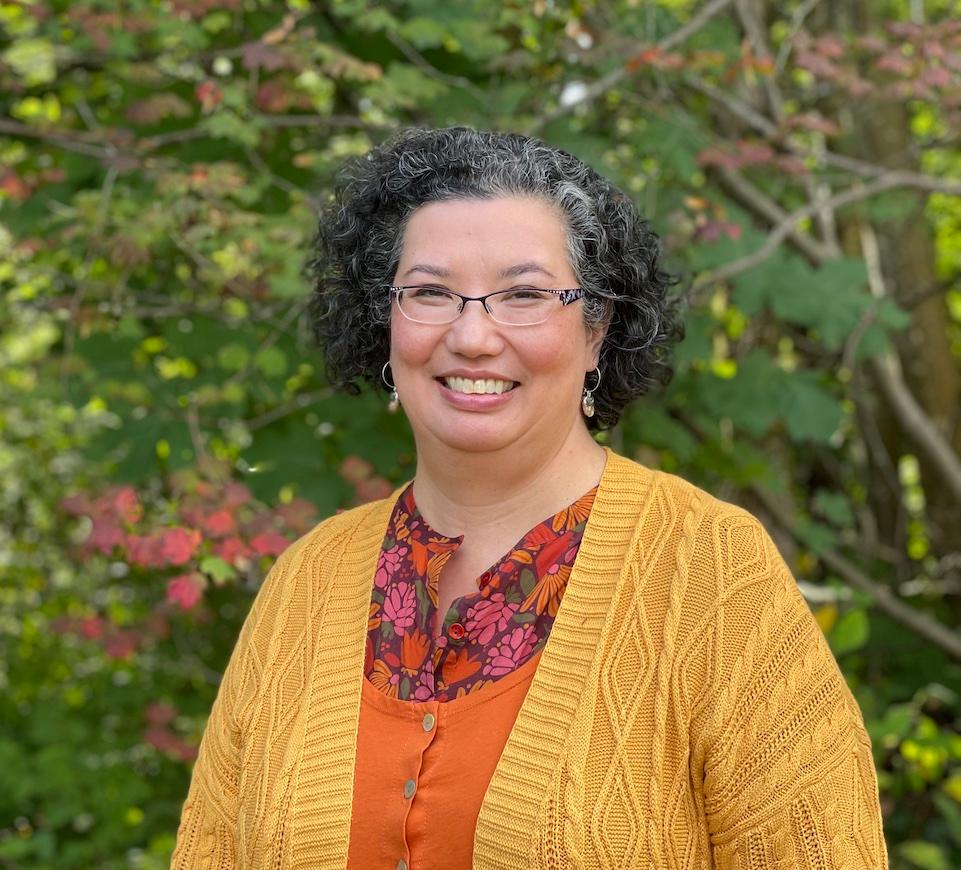 Half body shot of Sunni Nishimura. She is smiling at the camera with shoulder-length wavy black hair and glasses. She stands in front of a backdrop of autumn trees. 