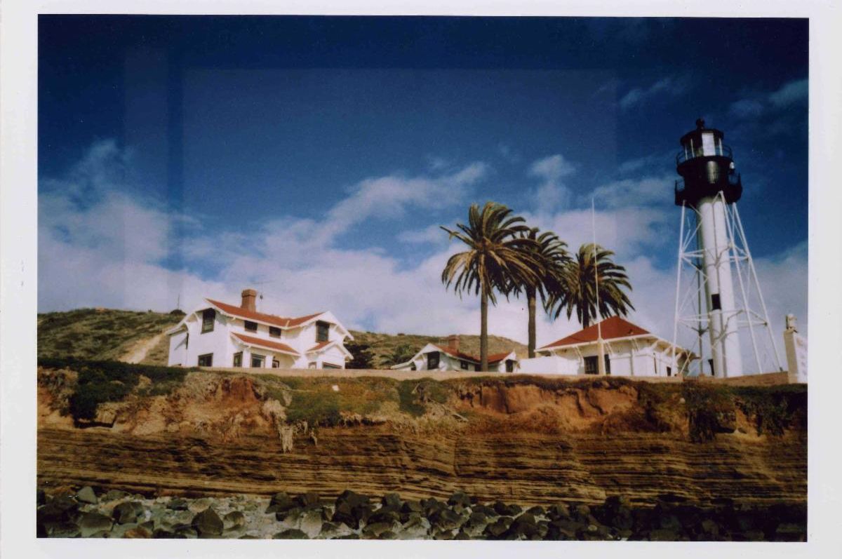 Polaroid of New Point Loma Lighthouse, as seen from shore, with its houses and tower. Two palm trees can be seen against a bright blue cloudy sky. 