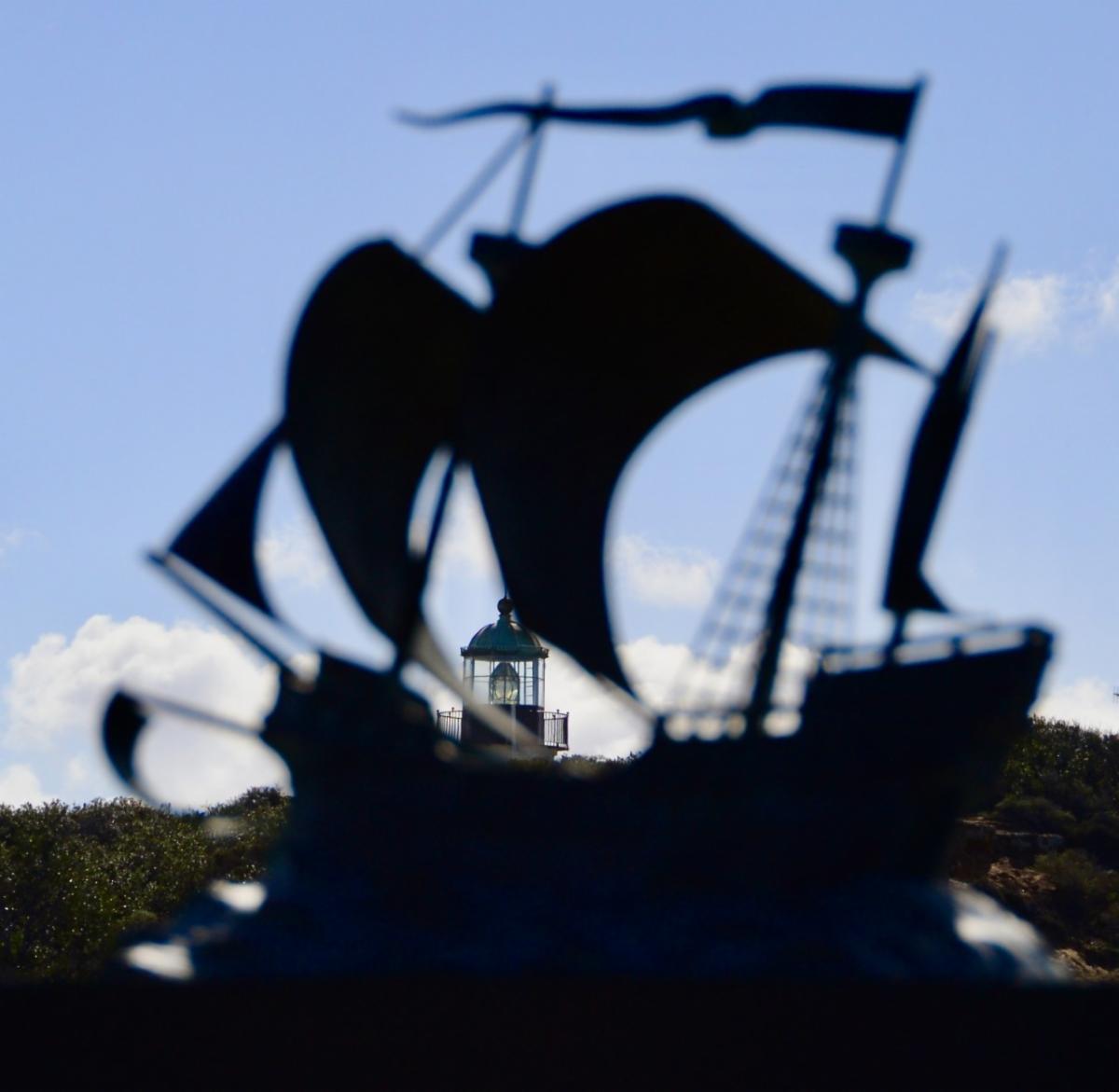 In the forefront is a small, out-of-focus statue of a 16th-century galleon. Captured perfectly between the sails, in the distant background, is the dark outline of the Old Point Loma Lighthouse against a blue sky.  