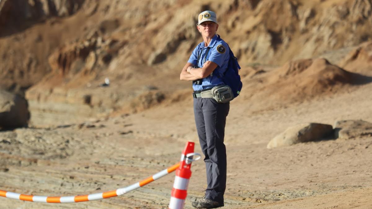 VIP Katrina stands tall in front of sandstone cliffs, with a small smile for the camera, as she safeguards visitors from a sensitive collapse area.