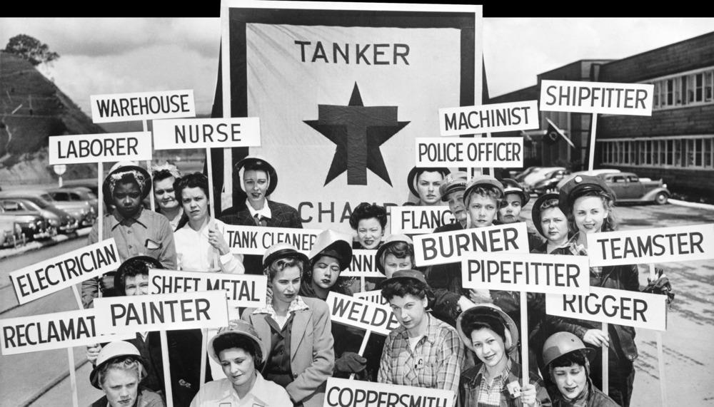 Black & white photo of women shipbuilders during WWII, holding signs listing their specific roles (like electrician, machinist, shipfitter, etc.).