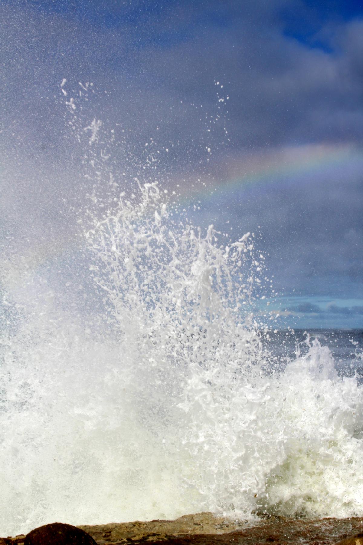  A massive wave crashes against the rocks, creating a rainbow over the white spray against the blue sky.