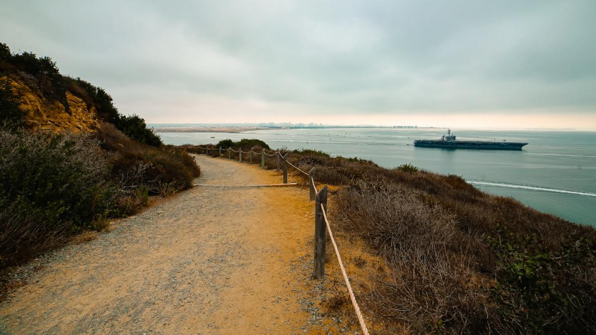 Bend of Bayside Trail overlooking the Bay, with a distant military carrier in the water under a cloudy horizon