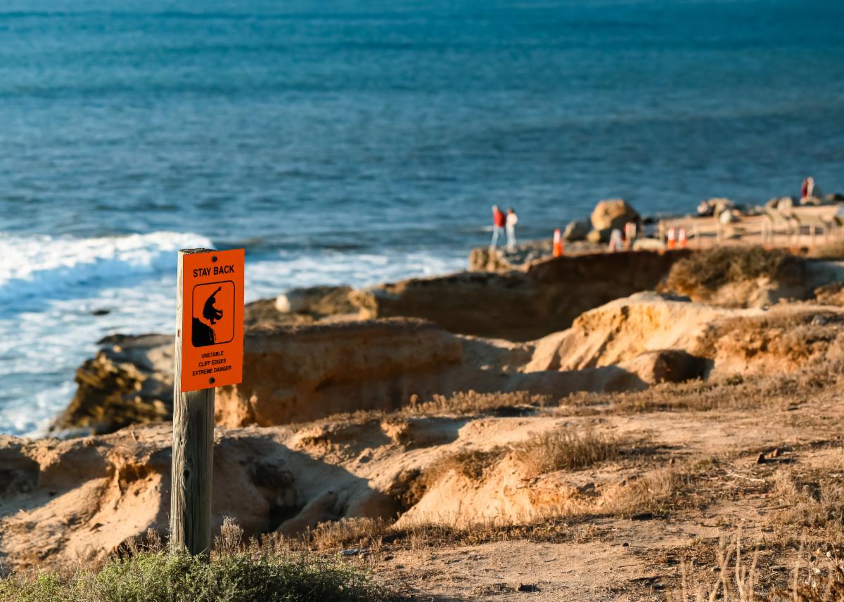 Unstable cliff sign at tidepool entrance. In the background, distant tourists observe the roaring waves by the cliffs. 