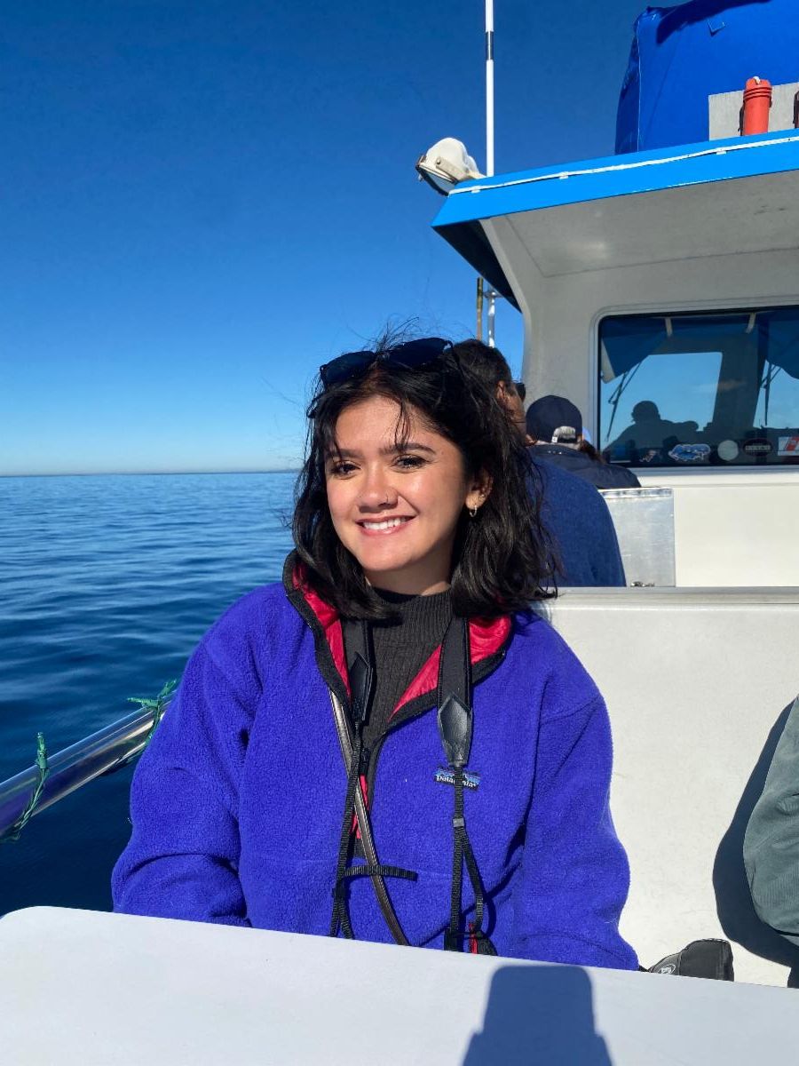 SiP Intern Kaelynn smiles as she sits on a small boat. The blue of her jacket matches the blue of the ocean and sky behind her.