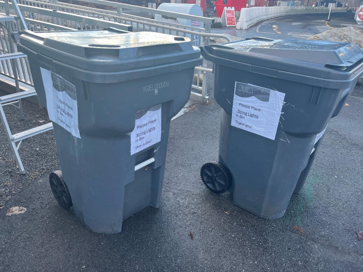 two large gray recycling bins sit on slightly damp pavement with printed paper signs taped to their sides