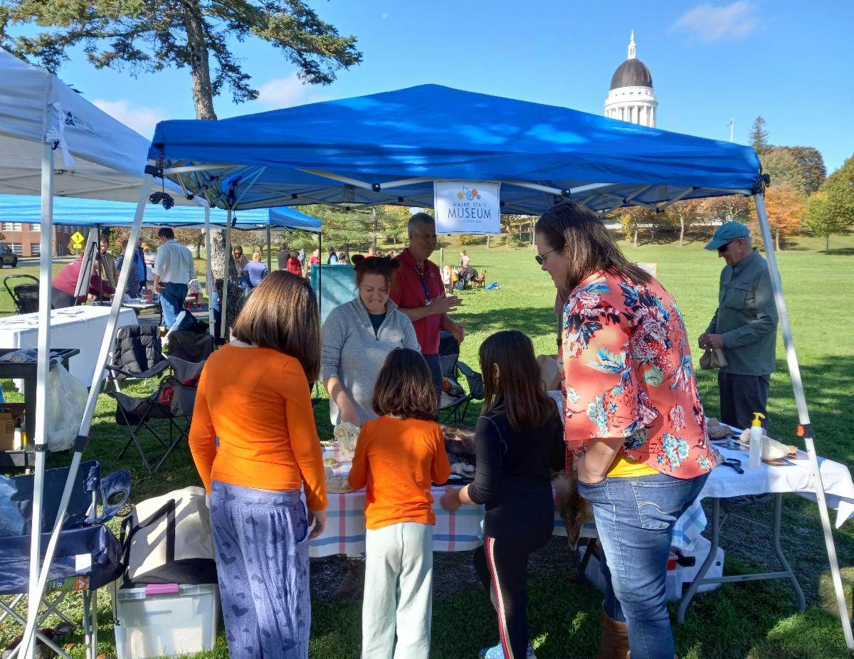 Table set up at an outdoor fair. A family talks with a museum educator. 