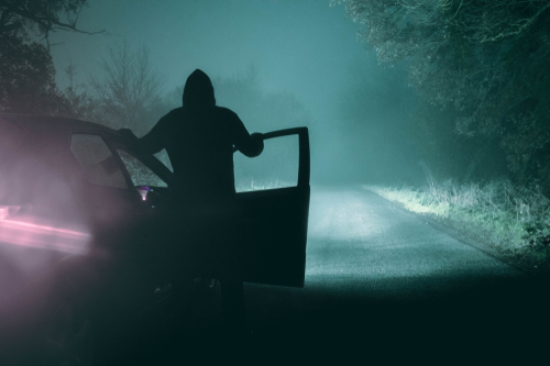 A lone_ hooded figure standing next to a car looking at an empty misty winter country road silhouetted at night by car headlights. With a cold_ grainy_ muted edit.