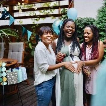A laughing pregnant woman with a friend on each side of her, each friend holding her hand over the pregnant woman's belly to form a heart