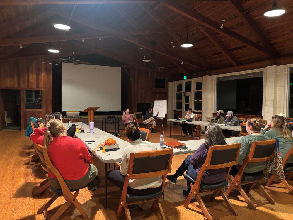 The program summit participants sitting around a table listening to a speaker at the front  of the room