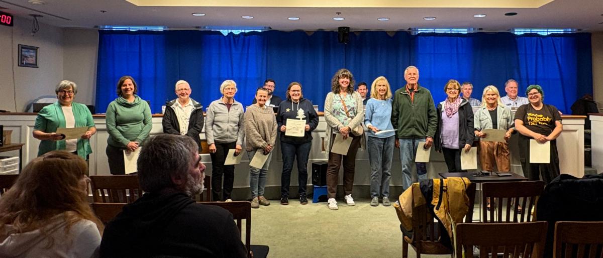 line of 20 or so people smiling facing camera standing in front of council dais