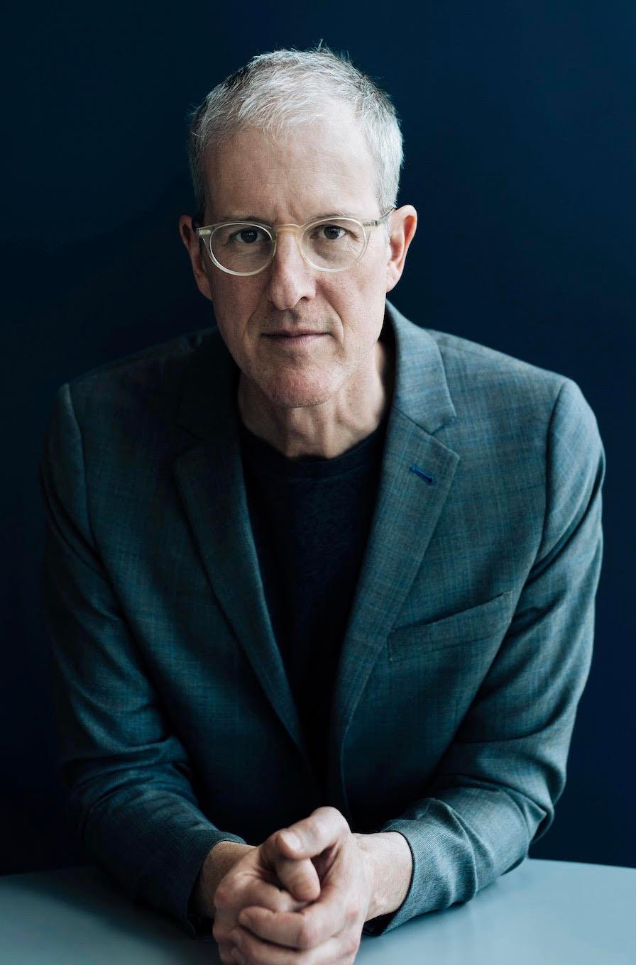 Bespeckled man in suit jacket staring at viewer, hands crossed in front of him