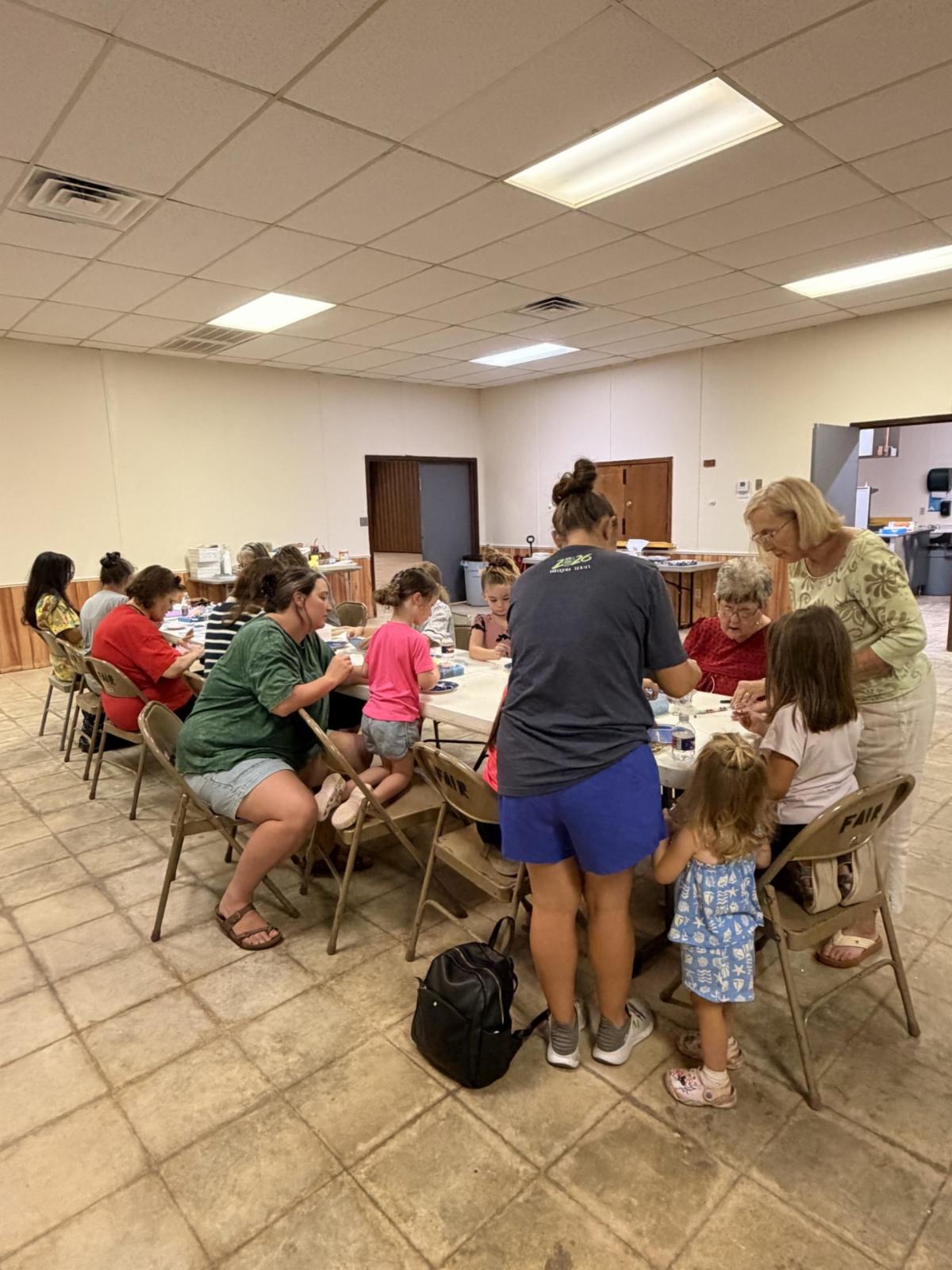 Community members of all ages gather around tables in a meeting room, participating in hands-on craft activities. Participants work together on projects while sharing supplies and instructions during a community workshop.