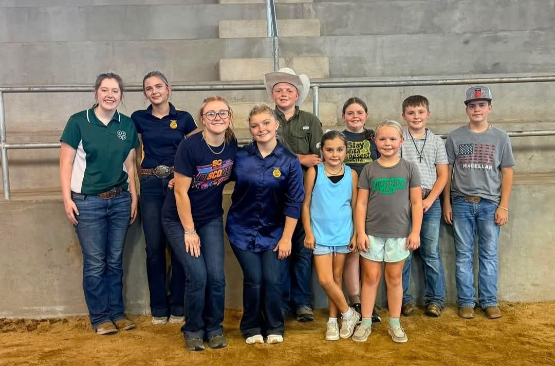 Group photo of ten youth standing together in a livestock show arena with a dirt floor and concrete bleachers in the background. Several participants are wearing 4-H and FFA shirts, jeans, boots, and western attire, including one youth wearing a white cowboy hat. Two younger children stand in the front row while the others are gathered behind them. The group is posed along the arena rail for a commemorative photo during the Blaine County Fair.