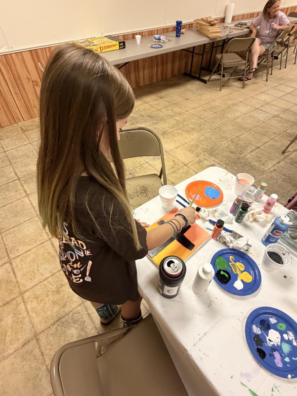 A youth participant paints a colorful canvas during an arts and crafts workshop. Paint bottles, brushes, palettes, and supplies are arranged on the table as the participant adds details to an artwork.