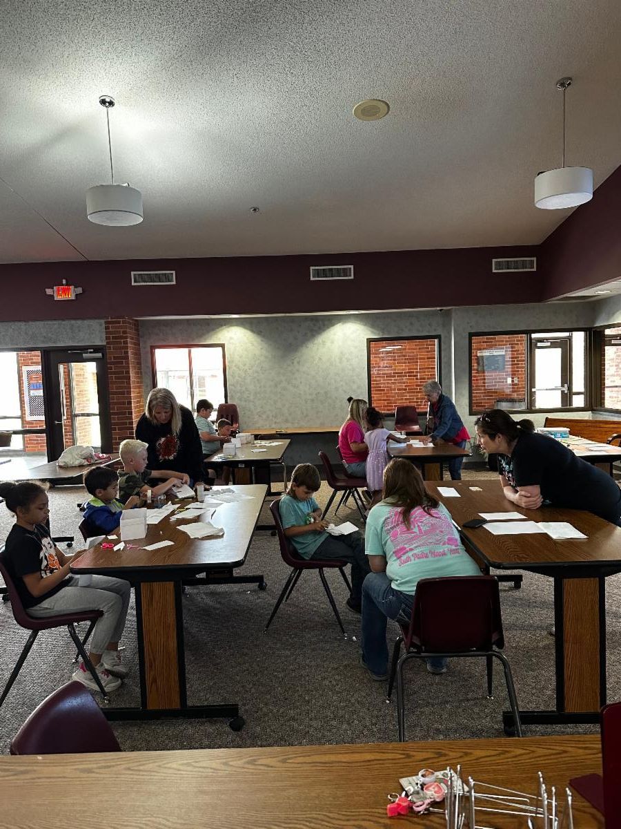 Children and adults sit at several tables in a large community room, cutting and assembling paper crafts. Some participants work individually while others receive help from adults standing beside them. The room features carpeted floors, brick and painted w