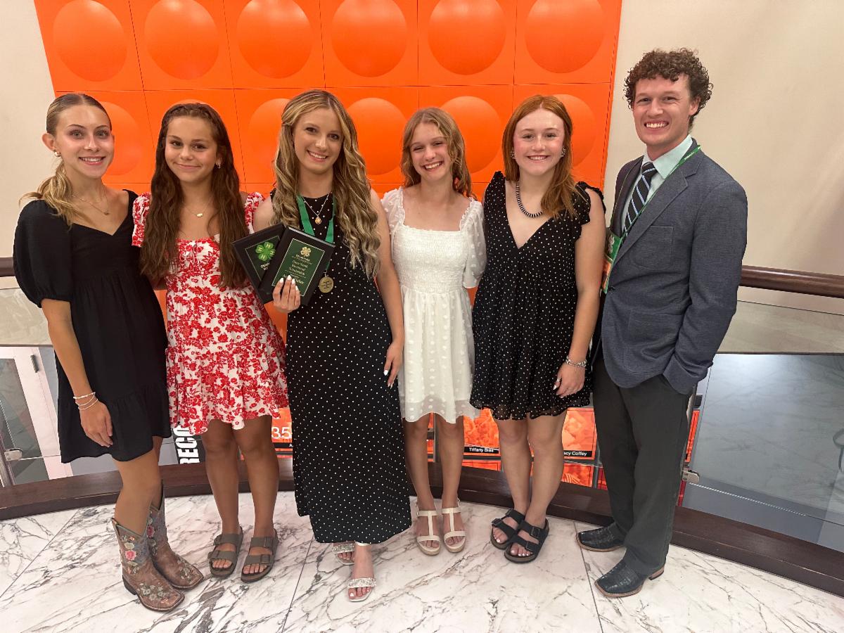 Six 4-H participants pose together at a recognition event, with one participant holding awards and a medallion, celebrating achievement, leadership, and involvement in 4-H.