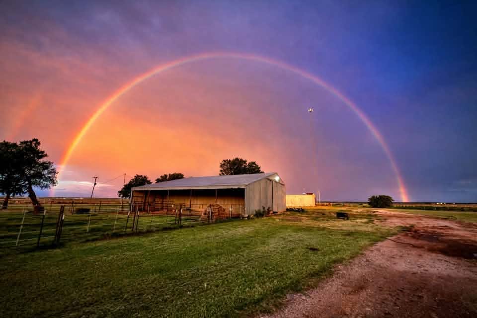 A wide rural landscape features a barn, fencing, grassy fields, and a gravel road under a colorful evening sky. A large rainbow spans nearly the entire width of the image, reaching from one horizon to the other. Shades of orange, pink, purple, and blue fill the sky, highlighting the countryside scene after a rain shower.
