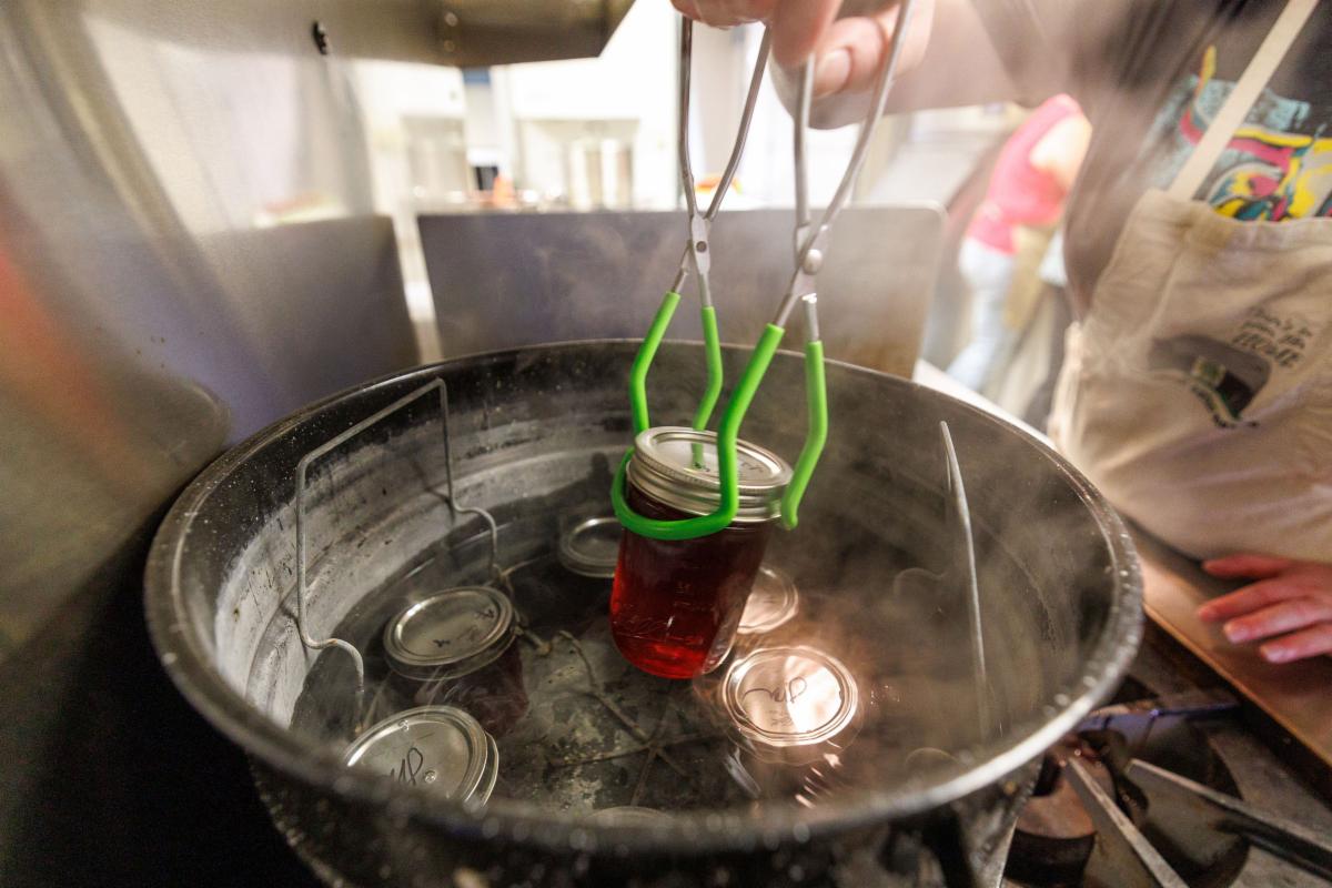 Close-up of a mason jar filled with red liquid being lifted out of a large metal pot of hot water using green-handled jar lifters, with steam rising and additional sealed jars visible inside the pot in a kitchen setting.
