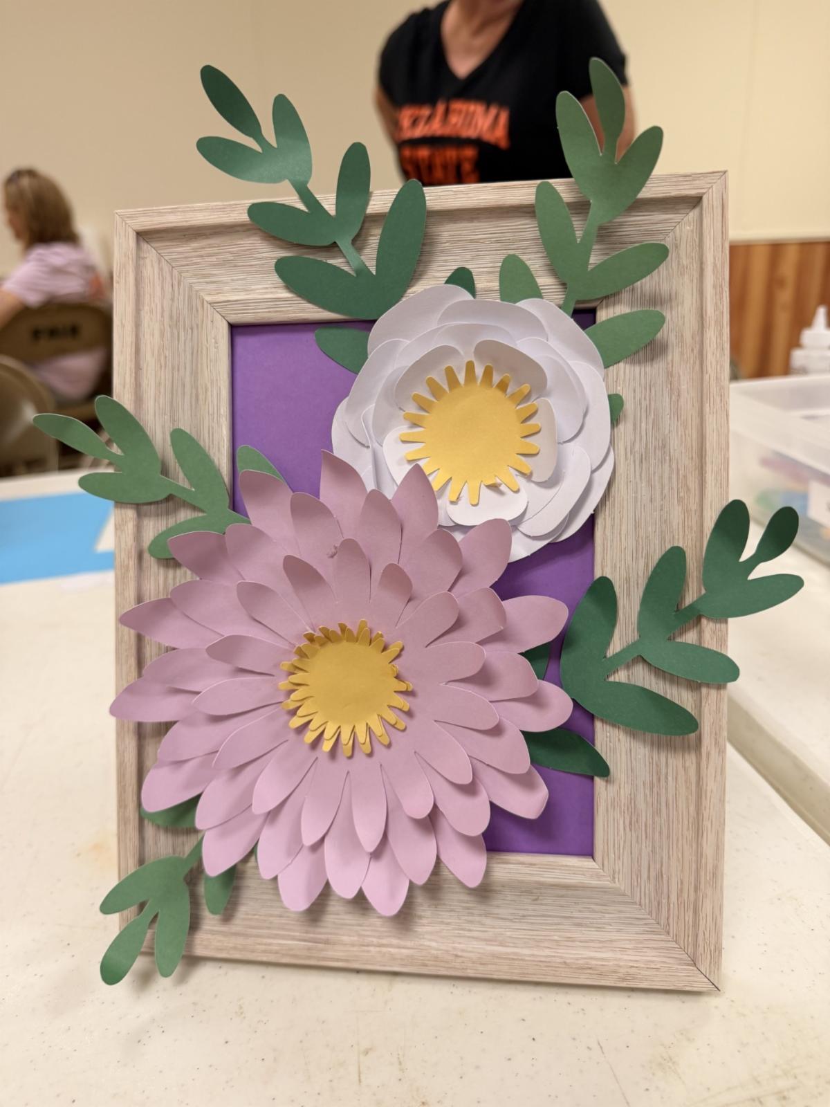 Framed paper flower artwork featuring layered pink and white flowers with yellow centers and green paper leaves arranged on a purple background. The handmade craft is displayed in a light wood frame.