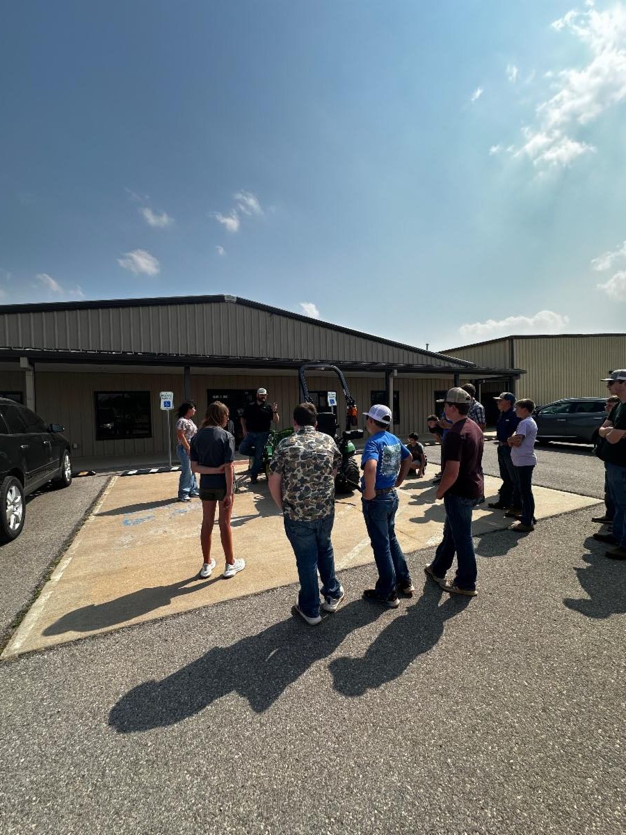 Group of youth and adults standing outside a metal building on a sunny day, gathered around a small green tractor as one person demonstrates or explains its use in a parking lot.