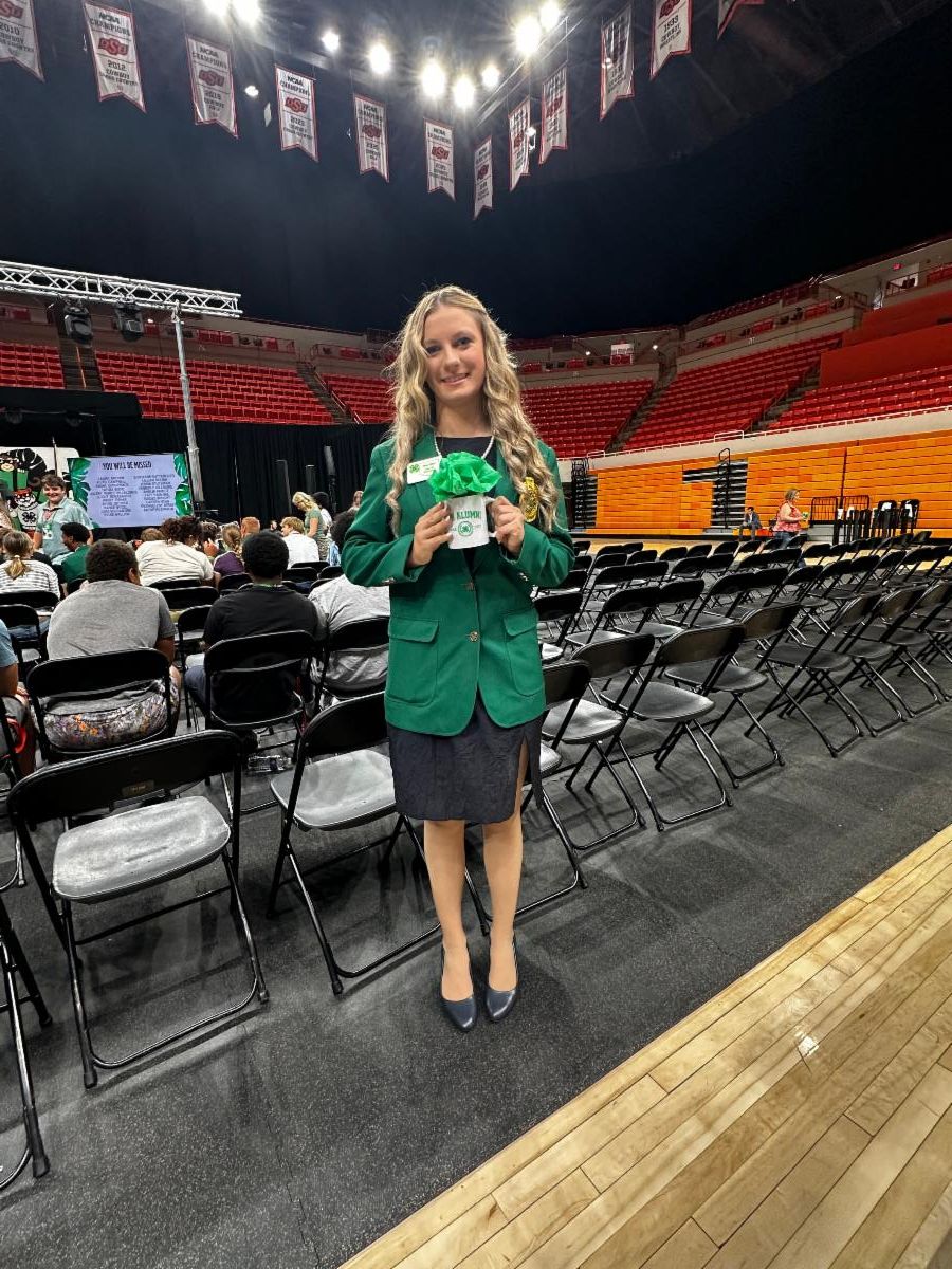 A 4-H participant wearing a green 4-H jacket stands in an arena during a statewide 4-H event, holding an alumni gift bag and celebrating leadership and achievement.