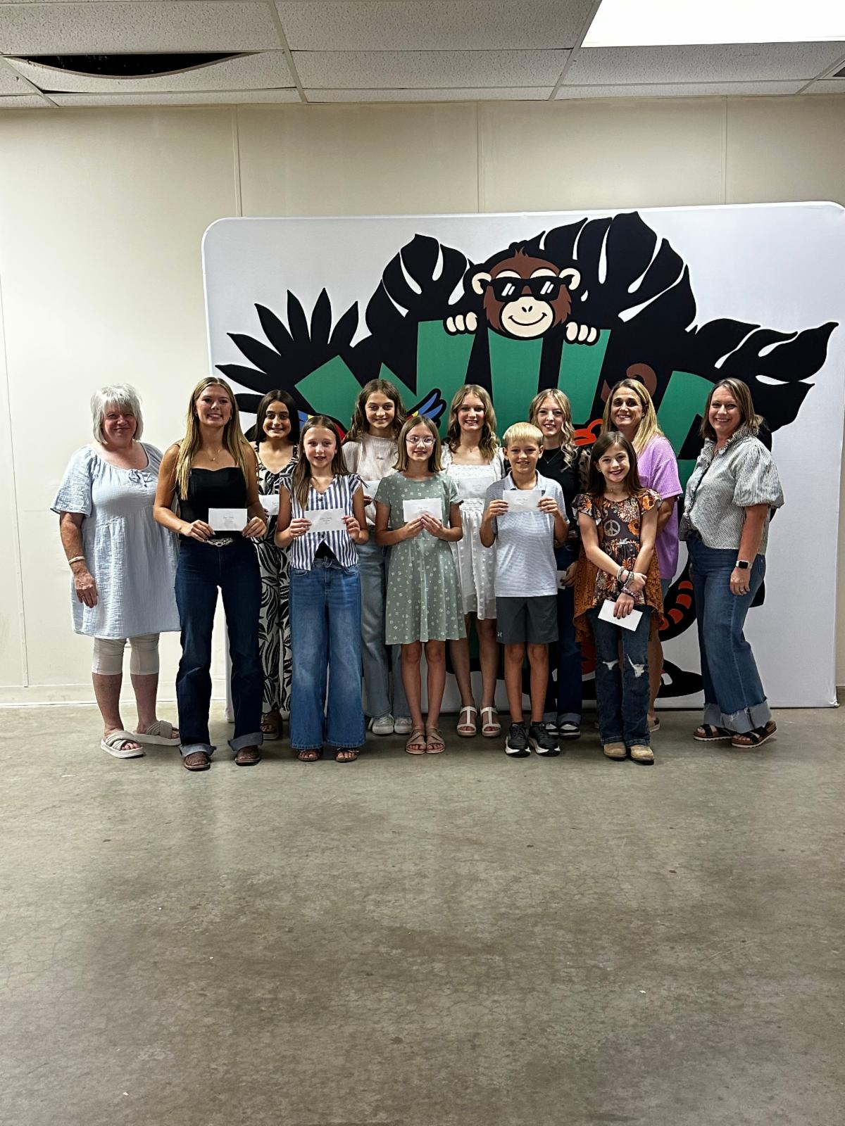 Okeene 4-H Club award winners pose with club leaders during the Blaine County 4-H Achievement Banquet. Several members hold recognition awards in front of a colorful 4-H backdrop.