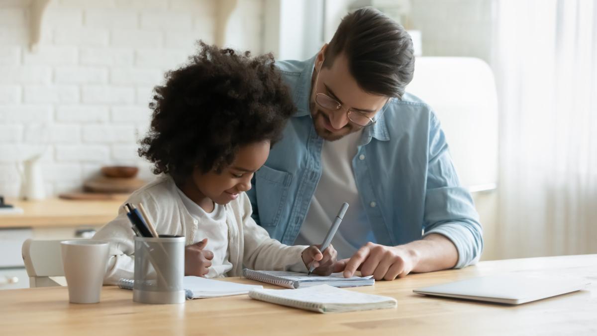 An adult helps a child with a writing activity at a table. Open notebooks, pencils, and a laptop sit nearby as the child practices writing with guidance and support.