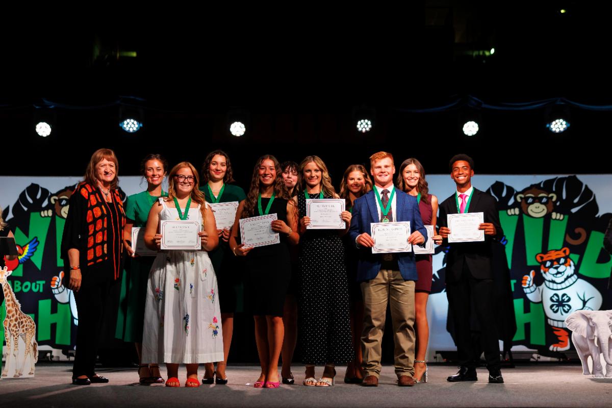 Avery Fisher stands with fellow award and scholarship recipients during a recognition ceremony at the 105th Oklahoma 4-H State Roundup. The group holds certificates on stage in front of 4-H-themed displays as awards are presented.