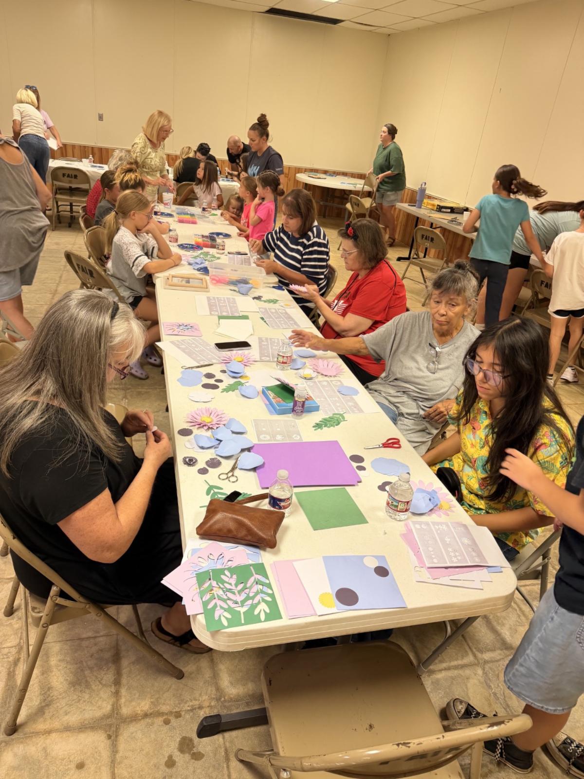 Community members of various ages sit around tables in a meeting room, creating paper flower crafts and cards using colorful paper, scissors, and craft supplies during a group activity.