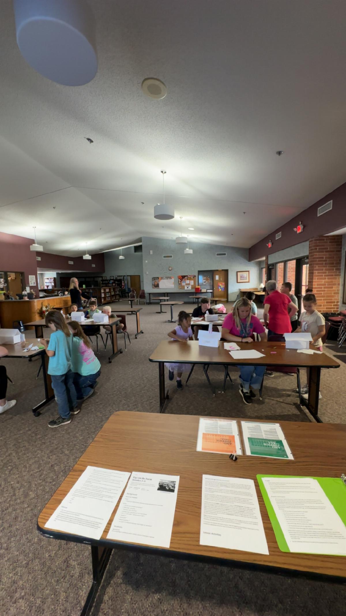 Children and adults sit at tables in a large, carpeted community room, working on paper activities and small white boxes, while informational handouts are spread on a table in the foreground; bookshelves, bulletin boards, and windows line the walls under a