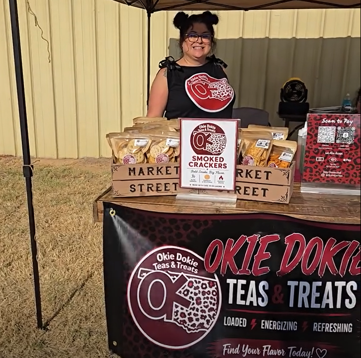 Vendor booth for Okie Dokie Teas & Treats at the Blaine County Fair. A vendor stands behind a display table under a canopy tent, with packaged snack products arranged in wooden crates. A sign in the center advertises smoked crackers, and a large banner across the front of the table displays the Okie Dokie Teas & Treats logo and branding. A QR code payment sign is visible on the right side of the booth, with a metal building and grassy fairgrounds area in the background.