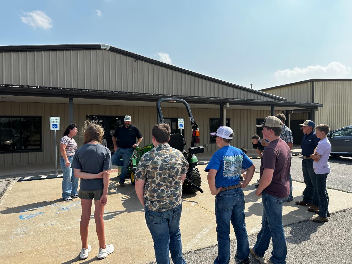 Group of youth standing outdoors in a parking lot beside a metal building, listening as an adult speaks and demonstrates equipment on a small green tractor during a hands-on learning activity.