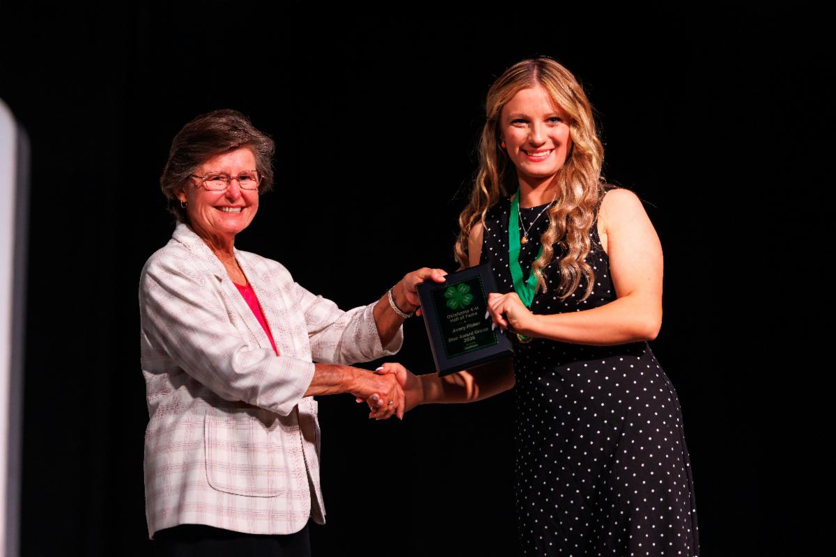 Avery Fisher receives an award plaque during the 105th Oklahoma 4-H State Roundup. Fisher and an award presenter pose on stage while holding the plaque and shaking hands.